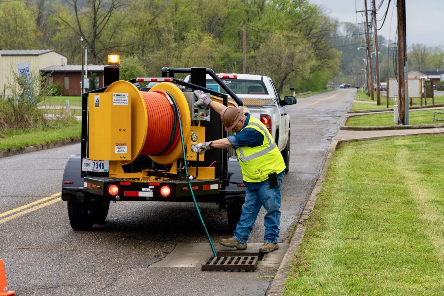 Worker operates O'Brien Sewer Jetters yellow trailer with hose reel to clear a roadside storm drain.